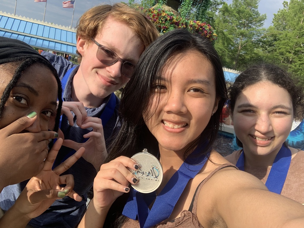 Four individuals smile at the camera. One holds a medal. Behind them, a building with glass windows and green trees.