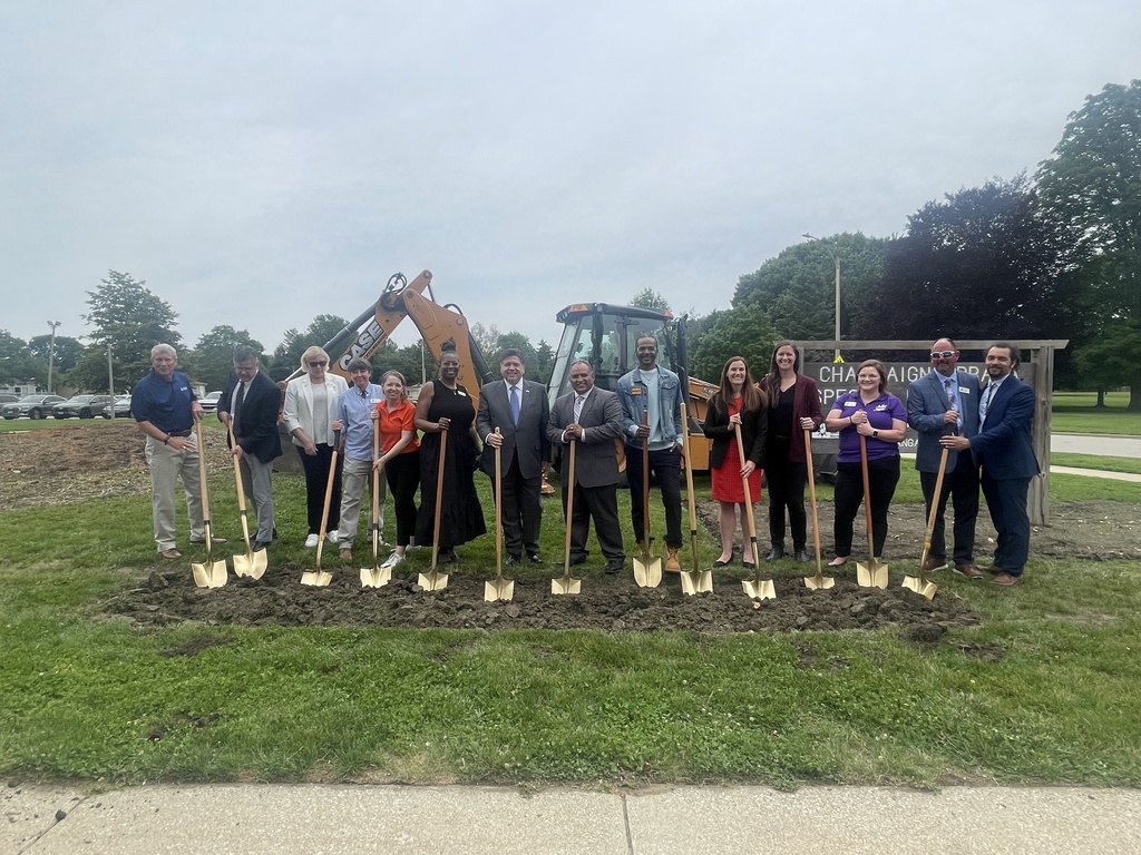 People hold shovels at a construction site, with a machinery vehicle in the background.