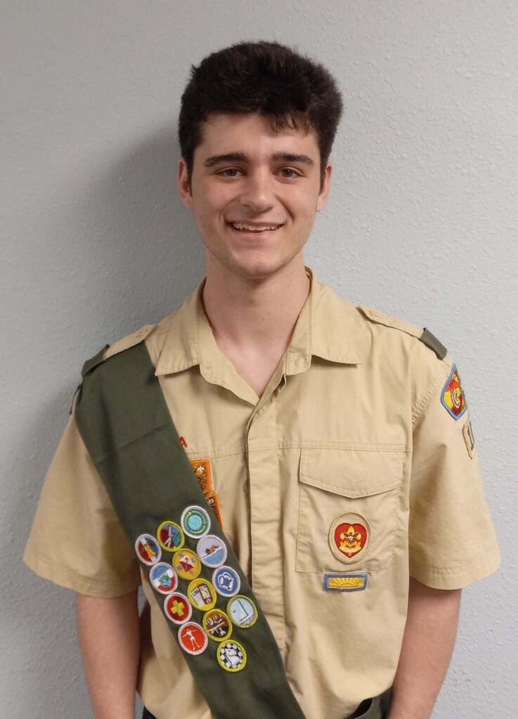 A smiling young man in a tan shirt and green sash with various badges stands against a white background.