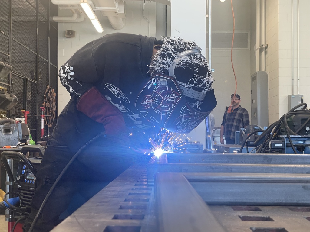 Person wearing protective gear welding metal in a workshop, with another person standing in the background.