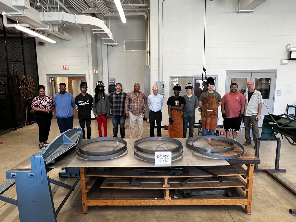 A group of people stands in a workshop with a table displaying large metal objects.
