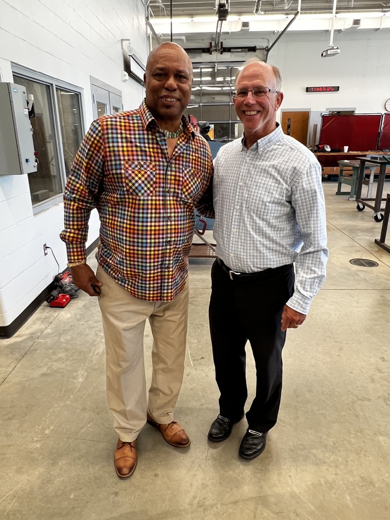 Two men standing in a workshop, one wearing glasses, a checkered shirt, and khaki pants, the other in a white shirt and dark pants.