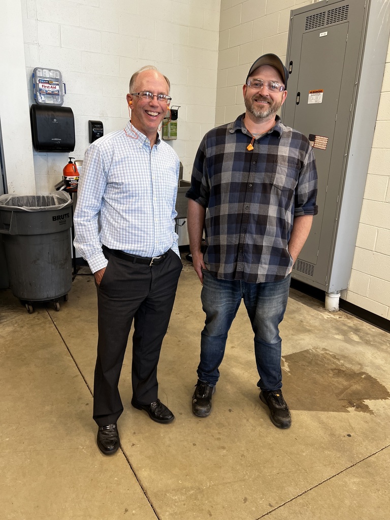 Two men are standing in a room with a fire extinguisher, trash bin, and metal lockers in the background.