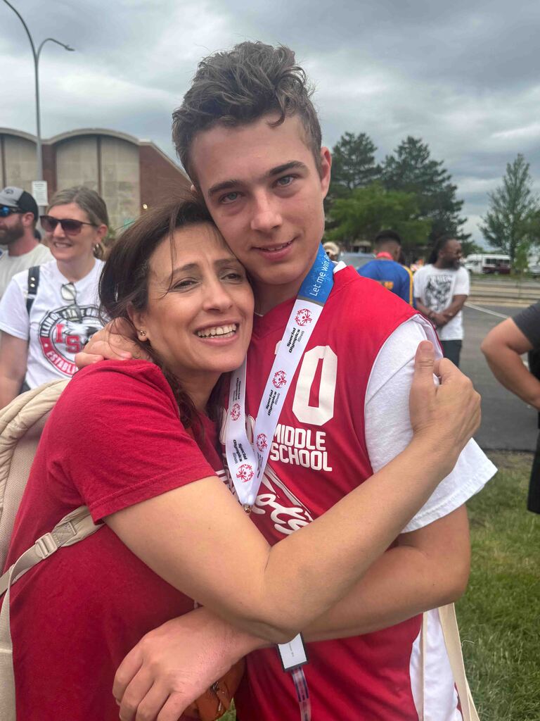 Woman in red and a boy in red and white sports jersey embrace while smiling. A large building is in the background.