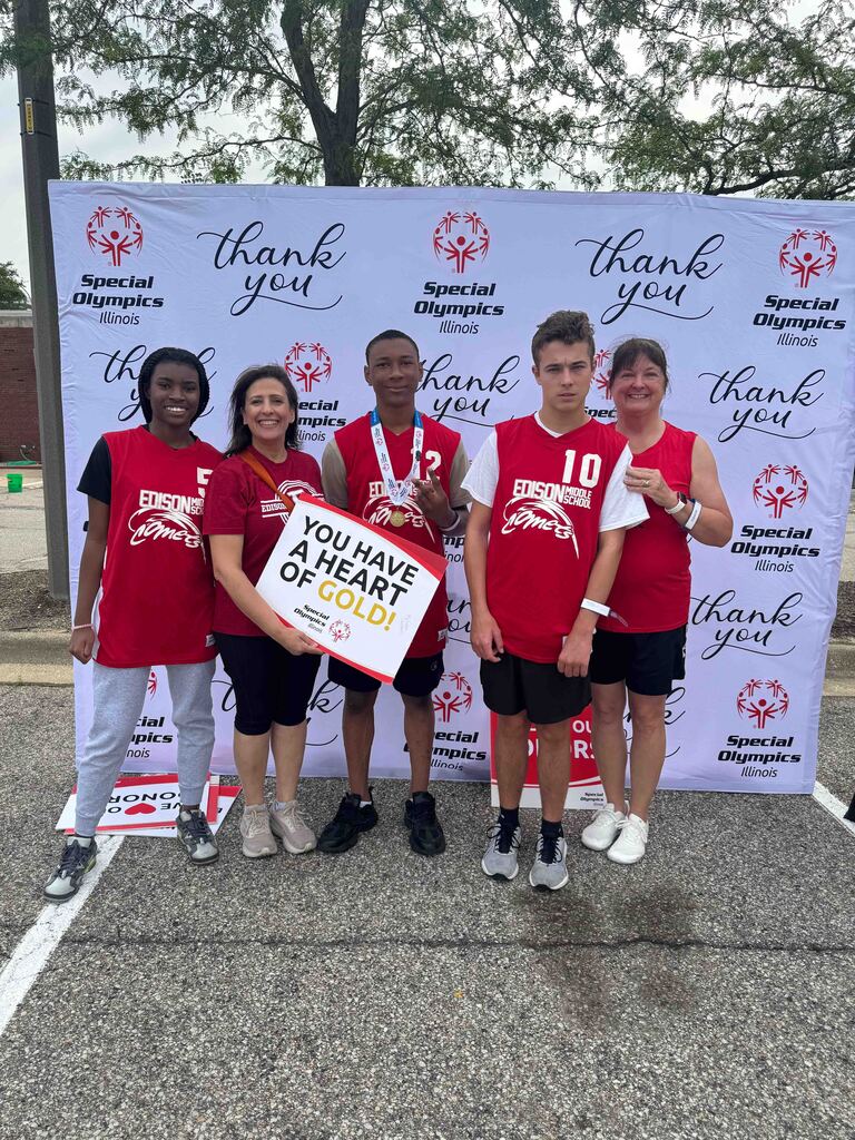 Six people pose for a photo at an event, wearing red shirts, holding a sign, and standing before a backdrop with a banner.