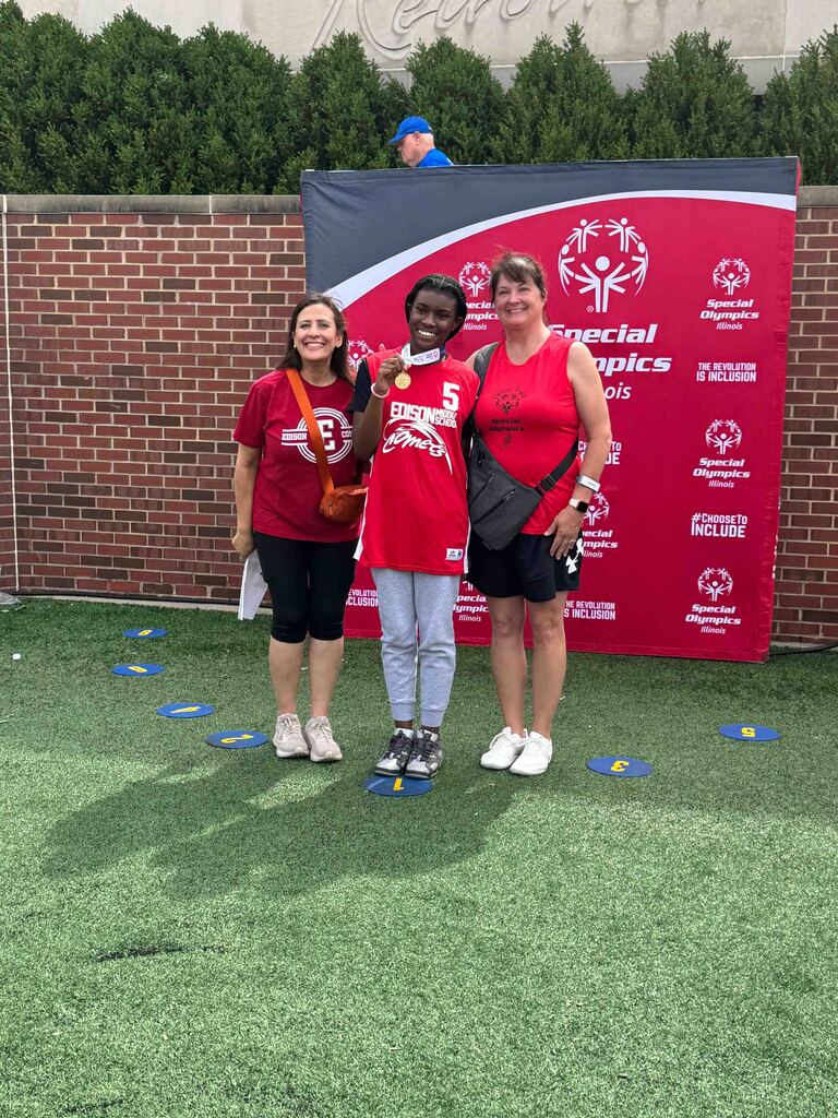 Three people in red shirts on a green field. One wears a medal. A red Special Olympics banner is behind them.