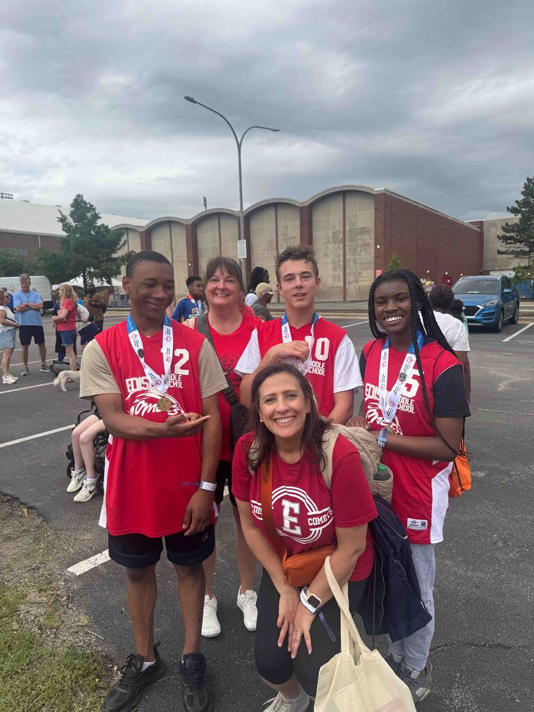 Five people in red athletic jerseys stand in a parking lot, smiling. Three have medals. A large building and trees are in the background.
