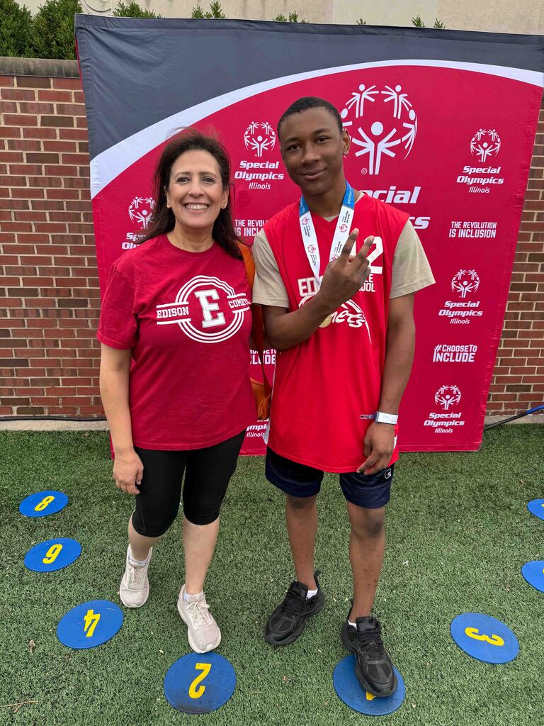 Two individuals in red shirts, one with a medal, stand on a green field with numbers. Behind them is a brick wall and a red banner.