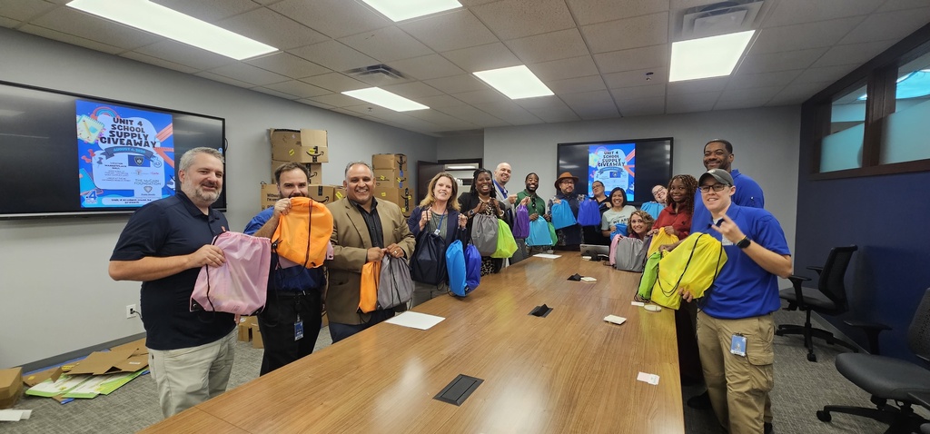 Group of adults holding bags, standing around a long table in an office with monitors on the wall.