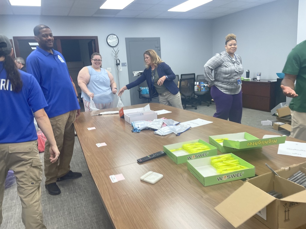 Six people standing around a table with boxes, papers, and a remote control on it.