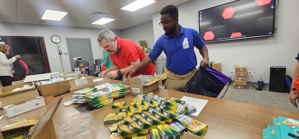 Multiple people sorting supplies in a room with tables, boxes, and a large screen on the wall.