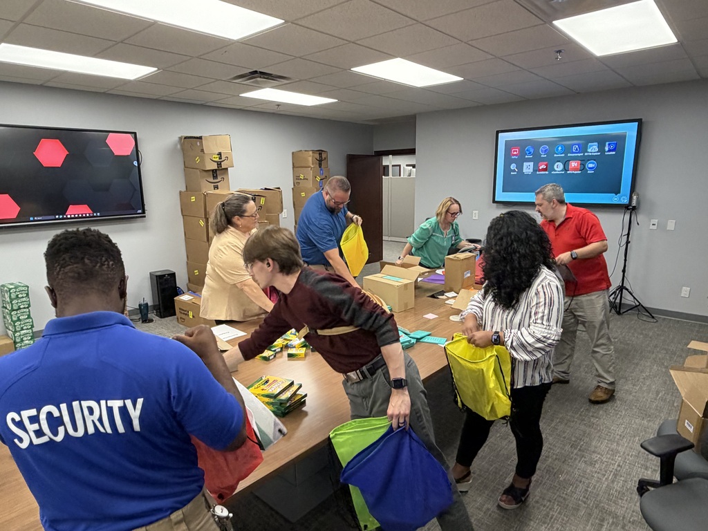 A group of people in a room with a table. Some hold bags. One person wears a blue shirt that says "SECURITY".