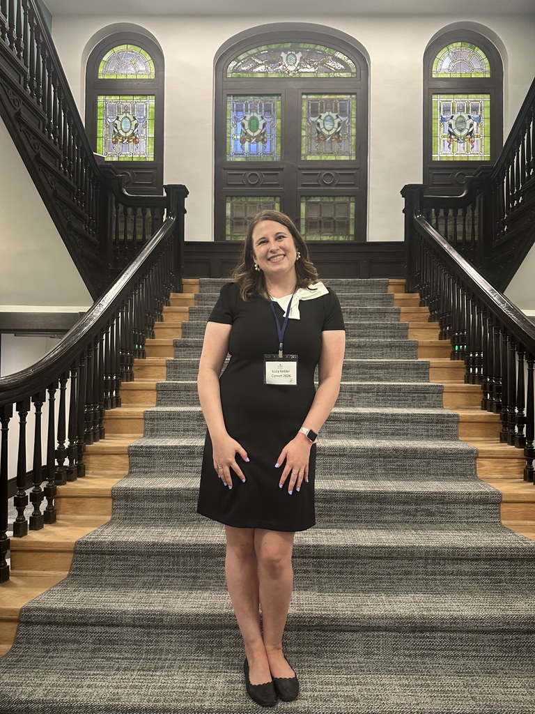 Lucy Kebler in a black dress stands on a carpeted staircase, hands on hips, with stained-glass windows behind.