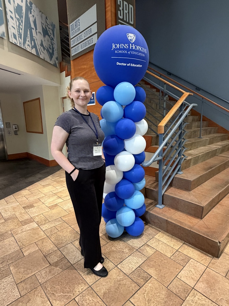 Taylor Kirtley stands next to a tall column of balloons. The column is blue and white.