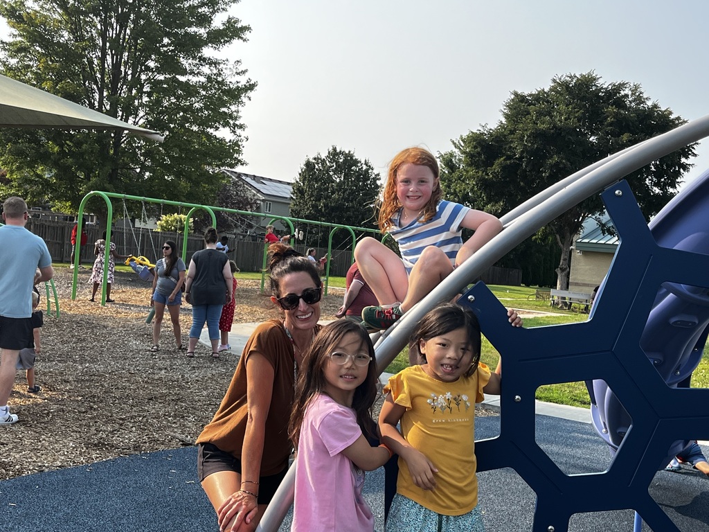 A woman and three children pose for a picture at a playground, surrounded by greenery and other people.