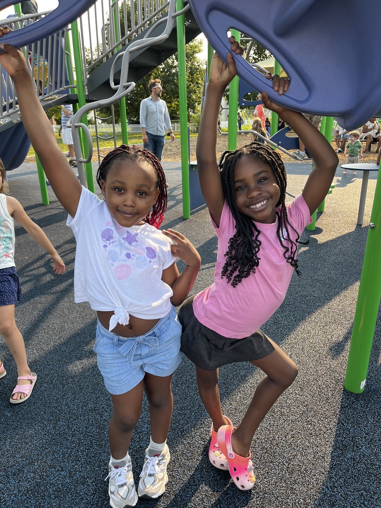 Two girls pose at a playground, smiling, with others nearby and a metal slide behind them.