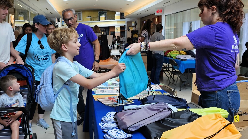 People gather in a hallway; a woman hands a boy a bag over a table with merchandise.