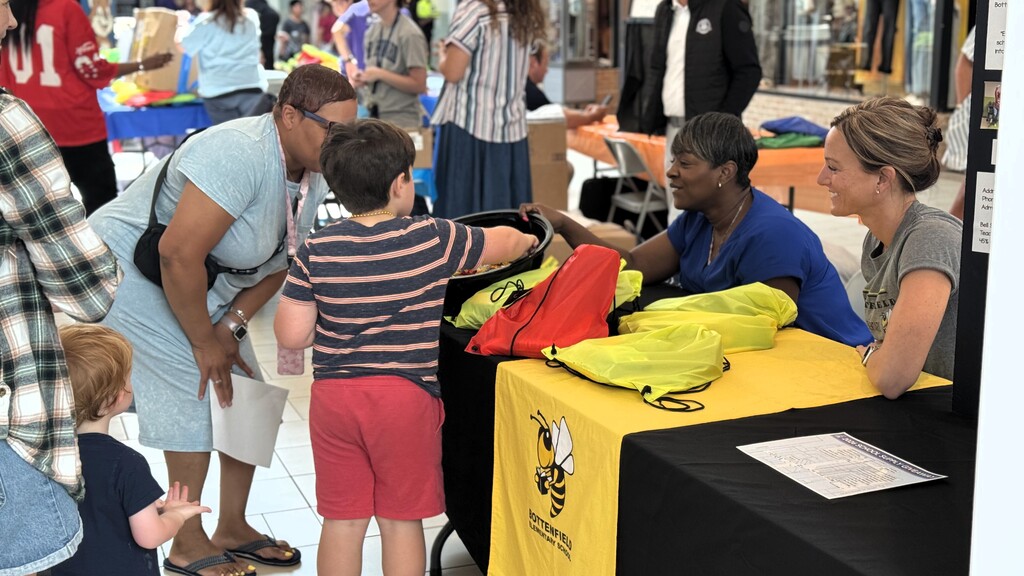 A group of people are standing around a table with a yellow tablecloth and a yellow logo.