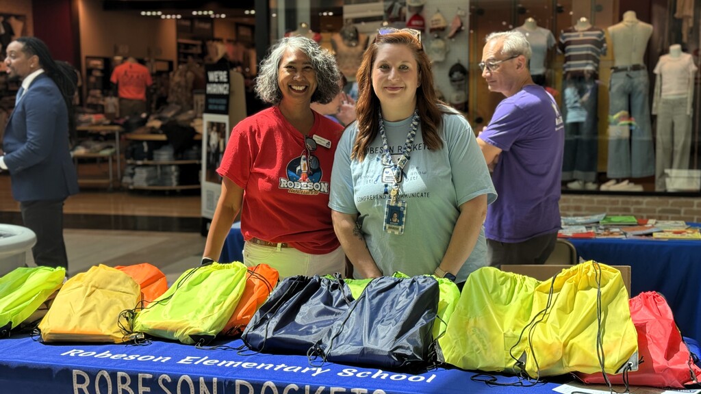 Two women stand behind a table with school supplies; one wears a red shirt, the other a blue shirt.