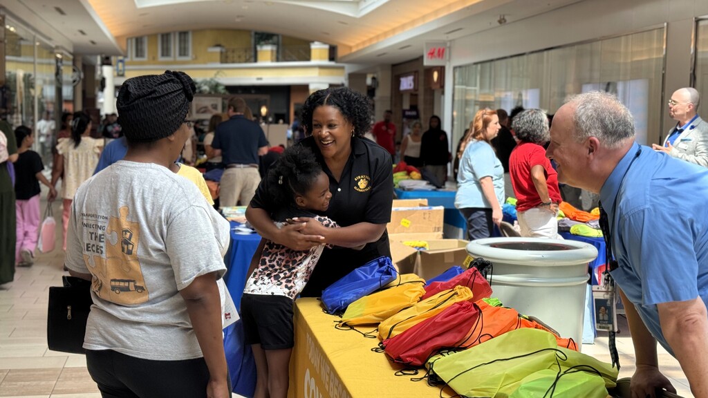 A crowd of people in a mall with several backpacks on a yellow table. A man and a woman are speaking.