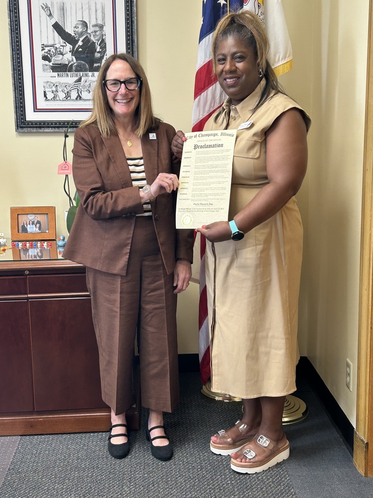 Two women in an office hold a certificate. One wears a brown suit, the other a beige dress. Behind them is a flag and a desk with a framed photo.