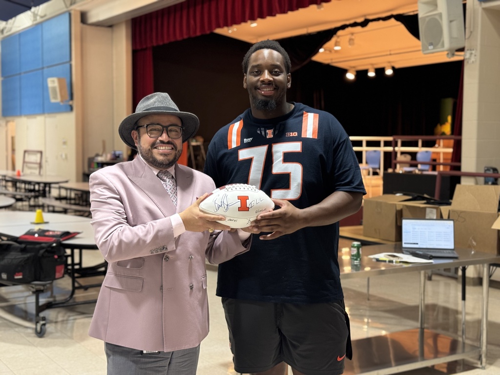 A man in a hat and suit holds a football while another man in a black jersey holds it too. Behind them is a stage and tables.