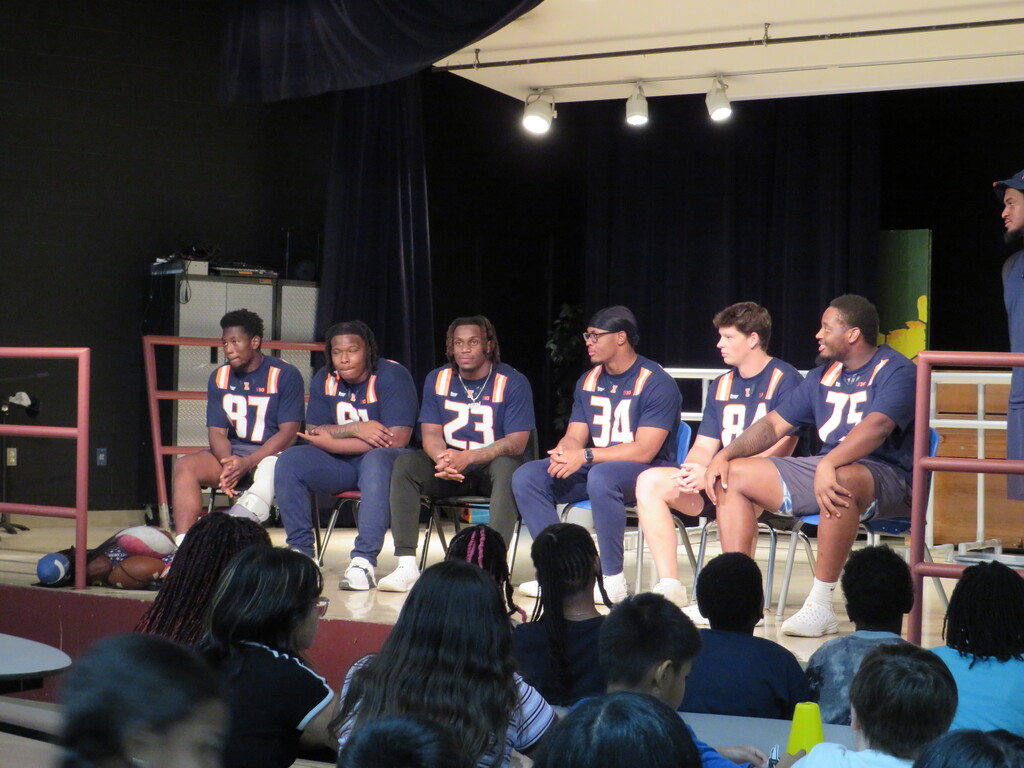 A group of athletes in blue uniforms sit on chairs on stage with numbers on the back. They face a crowd.