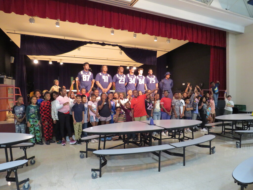 A group of people, including young children and adults, stands together on a stage with tables and chairs.