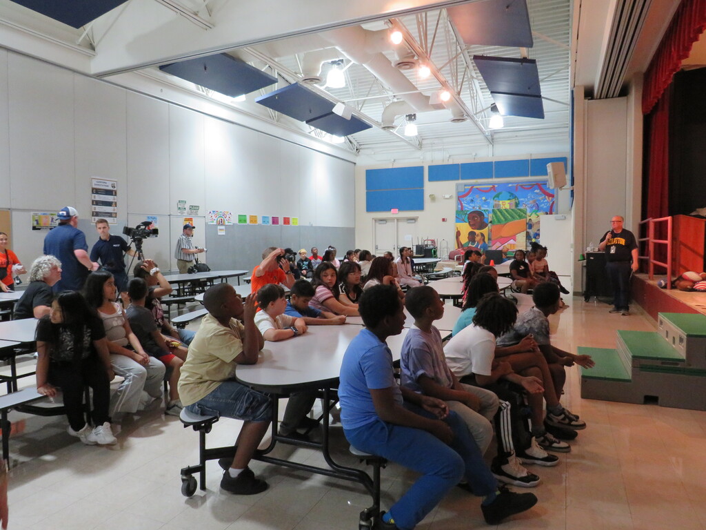 A group of children and adults sit at tables in a room with stage, curtains, and artwork.