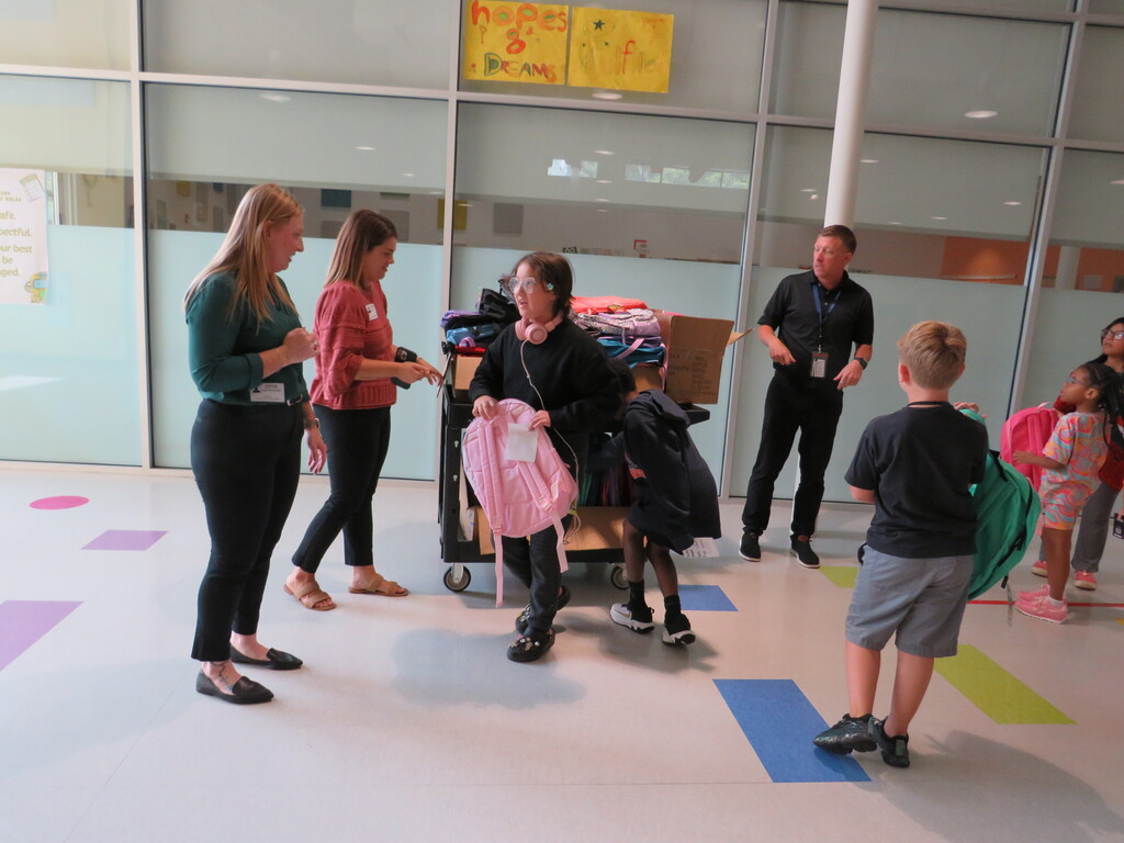Six people, including adults and children, in a room with glass walls. They are standing near a cart with bags.