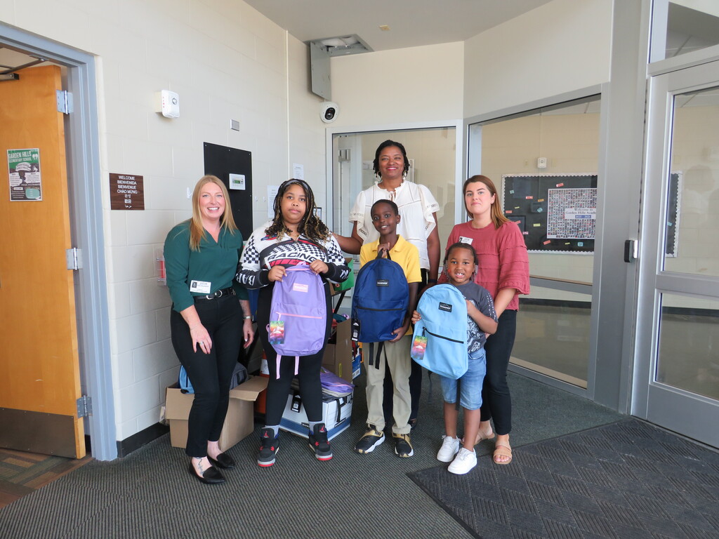 A group of adults and children stand together in a hallway, smiling. Each child holds a backpack.
