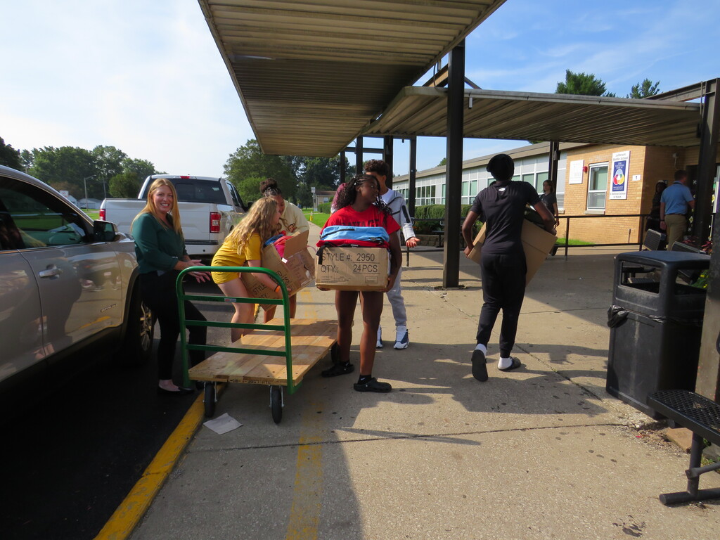 People move boxes on a cart under a canopy near parked vehicles. Some wear athletic gear.