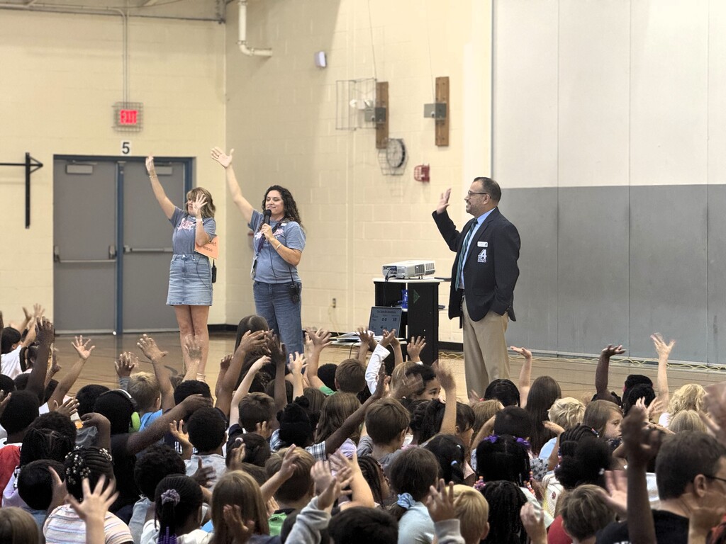 Three adults standing on a stage, one holding a microphone. Audience of children raising hands.