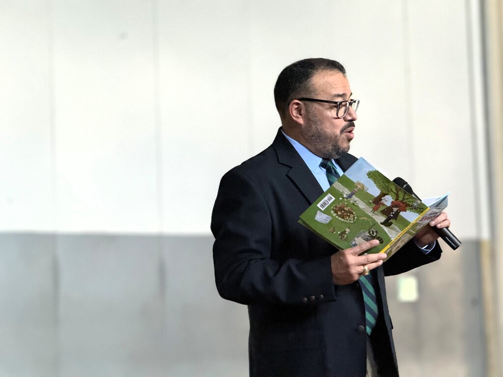 Superintendent Dr. Geovanny Poncy in a suit and tie holds an open book while speaking into a microphone.