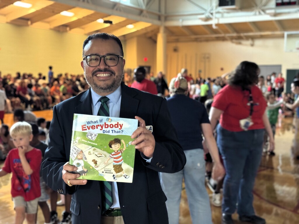 Superintendent Dr. Geovanny Ponce in a suit and glasses holds a book titled "What If Everybody Did That". Behind him, a crowd of people, including children, stands in a gymnasium.