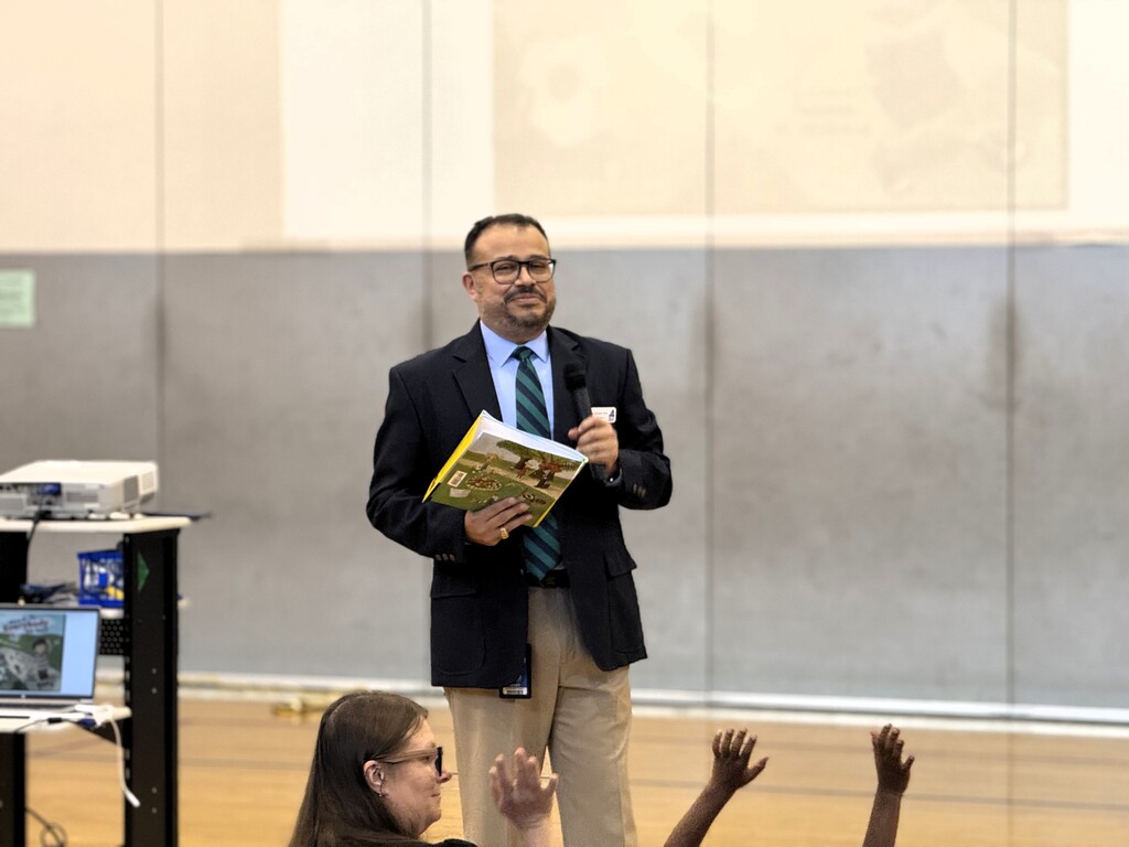 Superintendent Dr. Geovanny Ponce in a suit and tie stands in a room, holding a book. Children seated before him raise their hands.