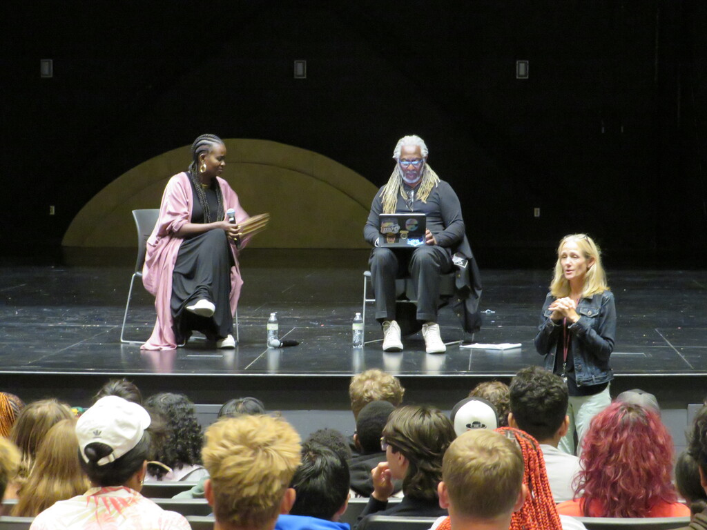 Three individuals on a stage in front of an audience, one seated with a laptop.