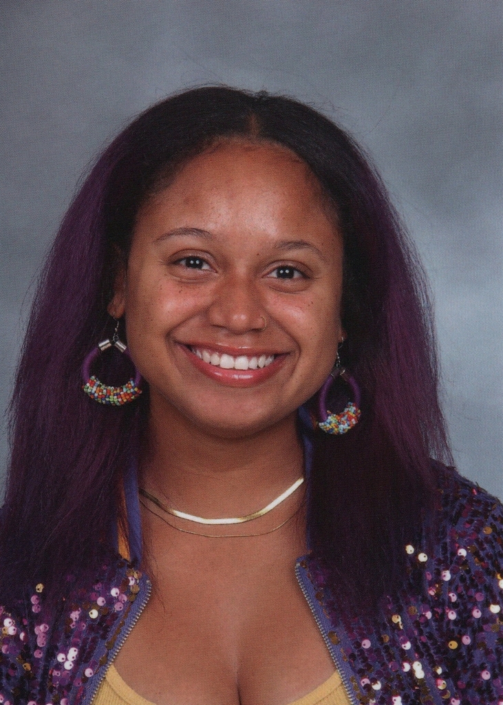 A woman with long purple hair wearing a yellow shirt and purple jacket. She smiles and wears colorful earrings.