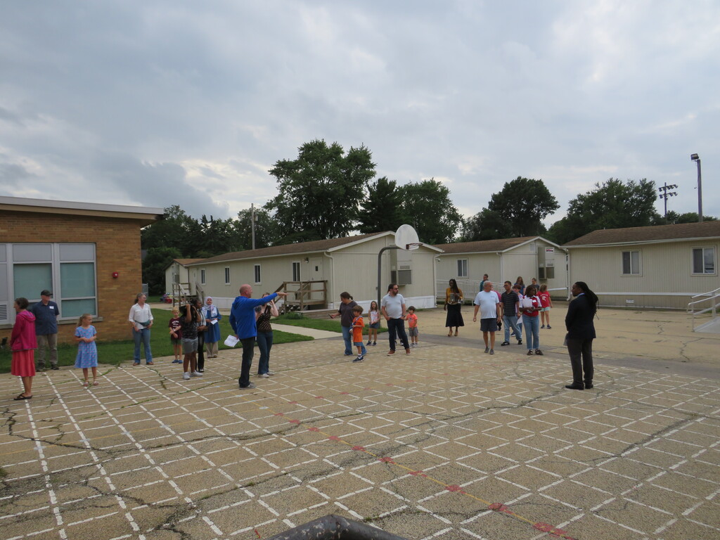 A group of people in various clothing and footwear are standing and walking on a brick floor. Behind them are houses with a basketball court.