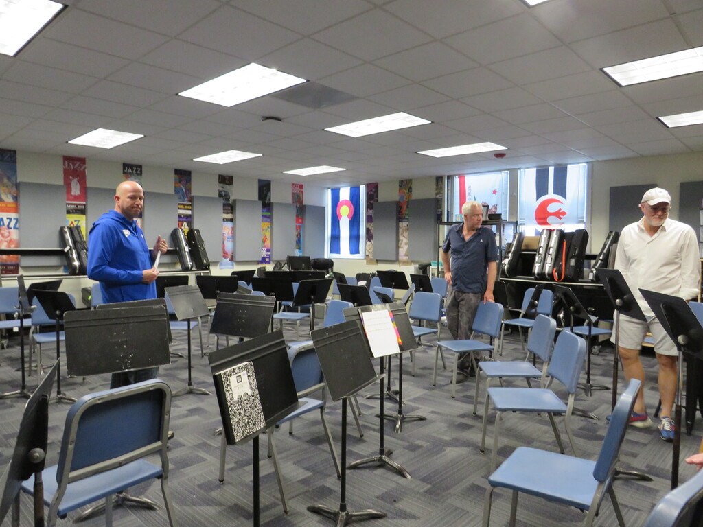 Three men in a room with musical instruments and chairs. One holds a piece of paper.