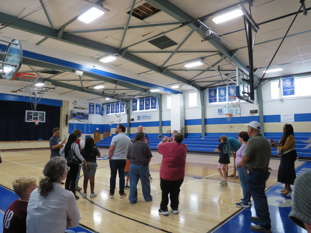 Several people gather inside a gymnasium. Blue bleachers are on one side. An elevated basketball hoop is on the opposite side.