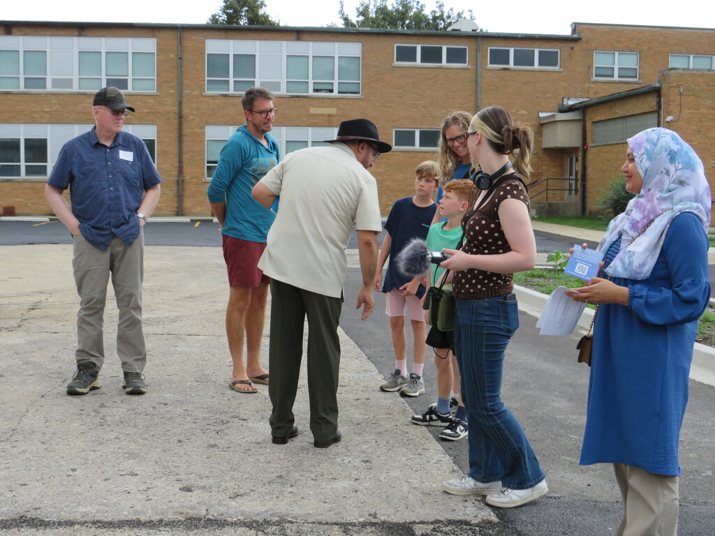 A group of people stands outside a brick building. One holds a microphone, and another holds a paper.