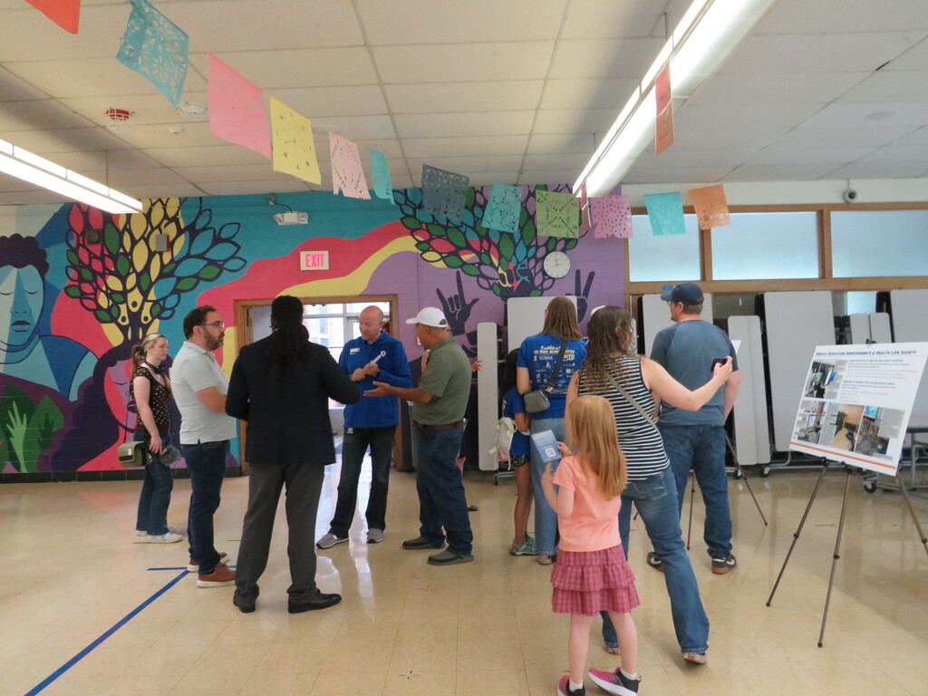 A room with a colorful mural features several people. A child stands in front of a board displaying plans.