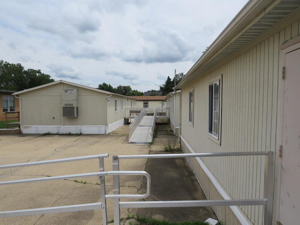 An exterior view of modular homes. A ramp connects the sidewalk to a door. Trees in the background.