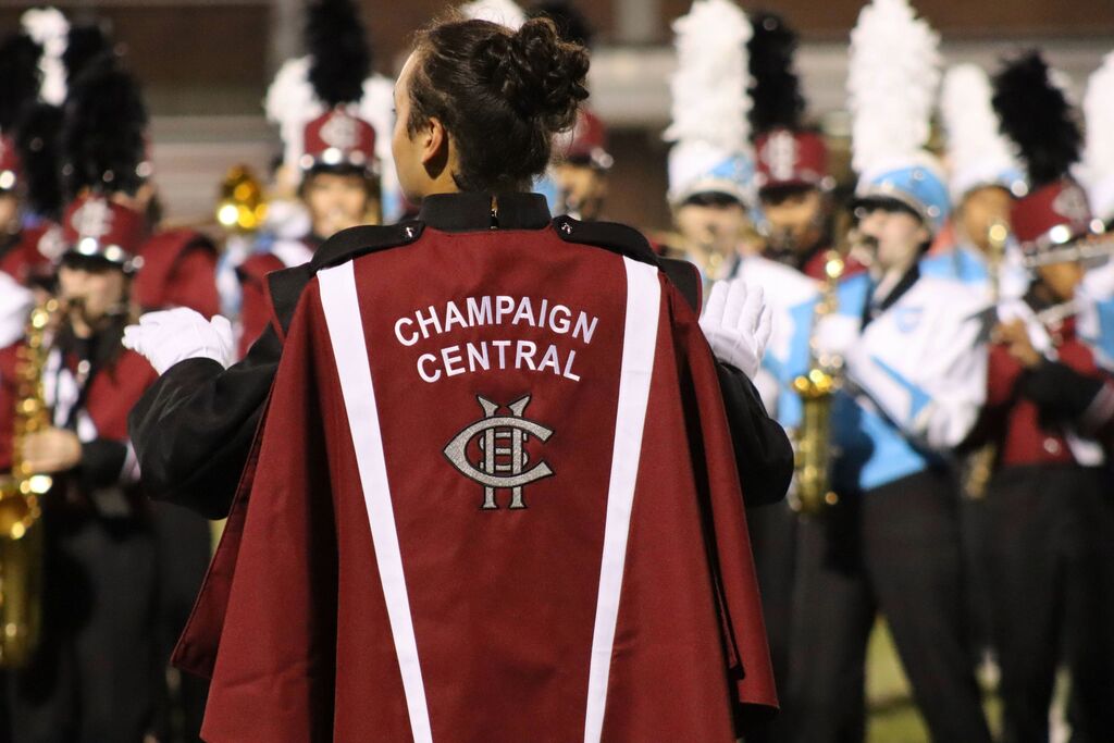 Central High School drum major wearing a maroon jacket with the words "Champaign Central" on it. Students in front of him are playing a variety of instruments including woodwind and brass.