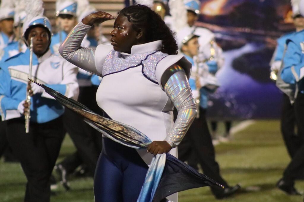A student performing with the Champaign Centennial High School color guard is wearing a white and blue uniform. She is holding a flag on a field, with band members in blue uniforms behind her on a field.