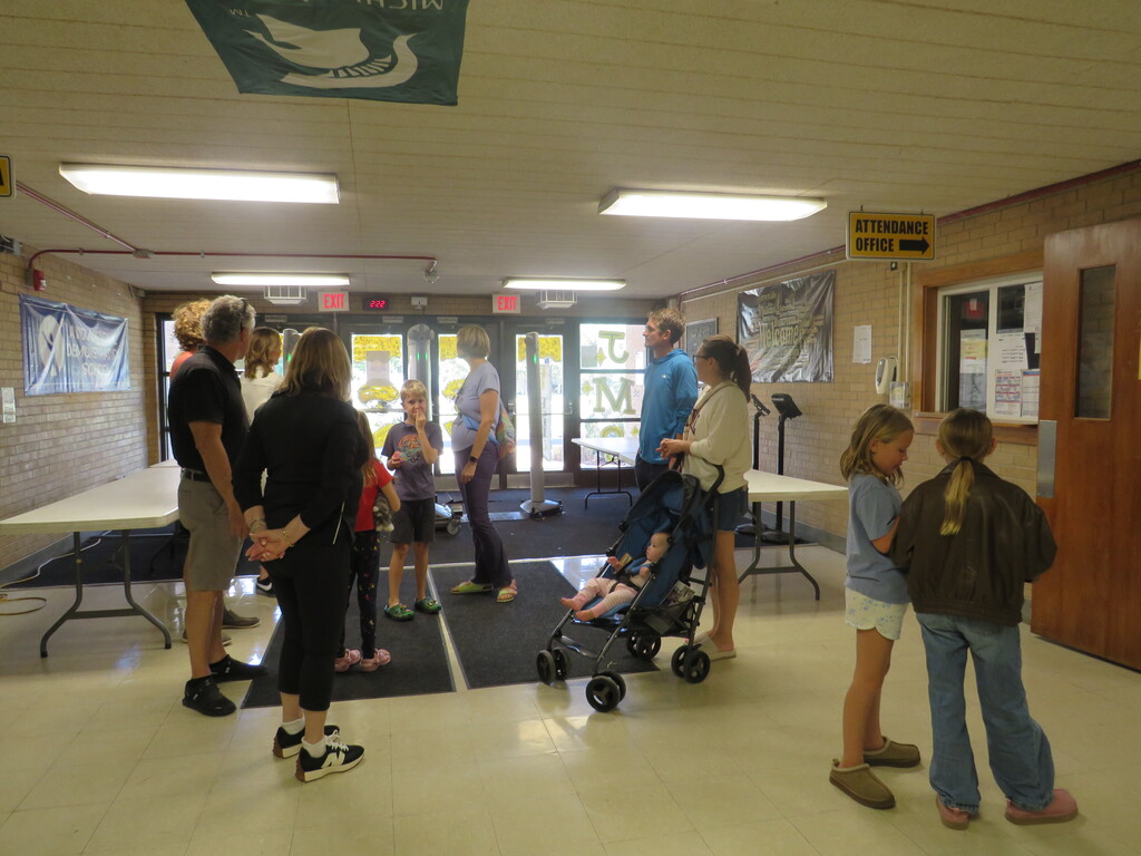 A group of adults and children in a room. Some are standing, some are sitting. A baby is in a stroller.