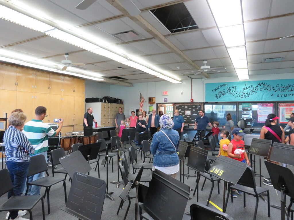 A room with music stands, flags, and people. Chairs are arranged in rows. A flag is on the wall.