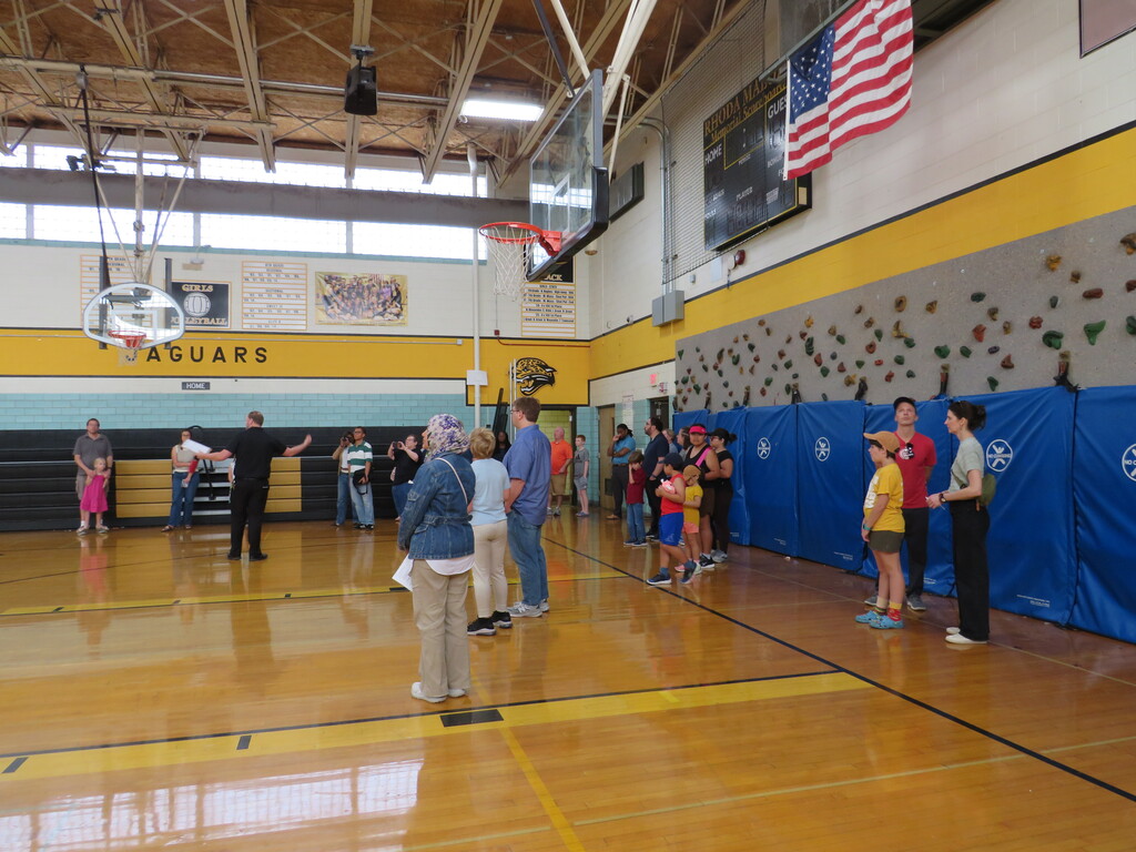 People in a gymnasium, some standing near a blue wall with climbing holds, others gathered near a basketball hoop.