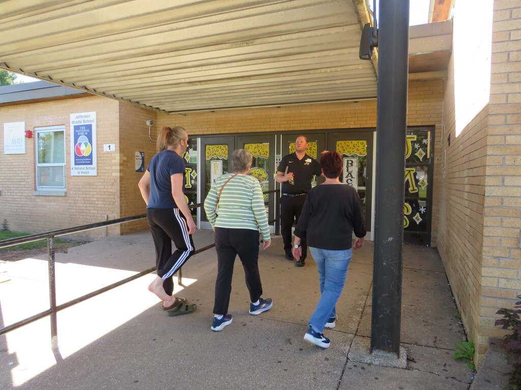 Four women in sneakers walk past a police officer. They are under a shelter in front of a building.